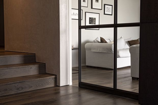 Interior view of a refined residential hallway or landing with dark timber flooring and matching timber stairs. A large black-framed, grid-panelled mirror reflects a pale upholstered sofa, cushions, and a gallery wall of framed artwork. The space has a warm neutral palette with strong black detailing and contemporary built-in architectural elements.