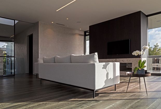 A refined contemporary living room with a low-profile light-grey sofa, dark timber television feature wall and integrated fireplace or media cabinetry. Full-height glazing brings in strong natural light, while dark timber flooring, a textured neutral wall, recessed ceiling lighting and a minimalist side table with an orchid create a calm, high-contrast interior.