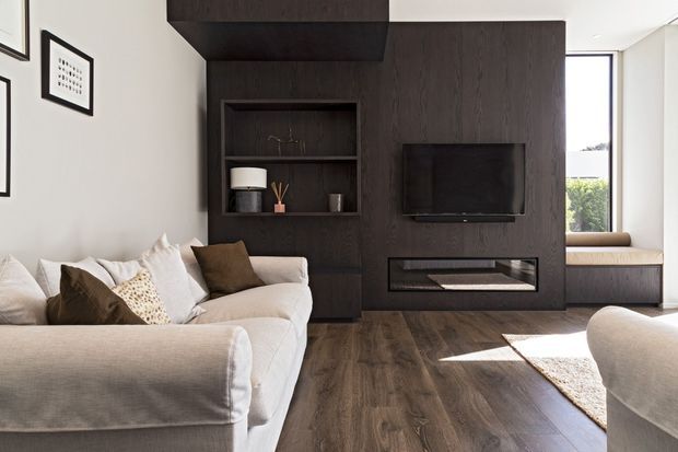 A refined contemporary living room with a large light upholstered sectional sofa facing a dark timber feature wall. The wall incorporates open shelving, a wall-mounted television, and a long linear built-in fireplace. Dark timber flooring, neutral furnishings, framed artwork, and a narrow window seat create a warm, balanced interior with strong contrast between the pale seating and dark feature joinery.