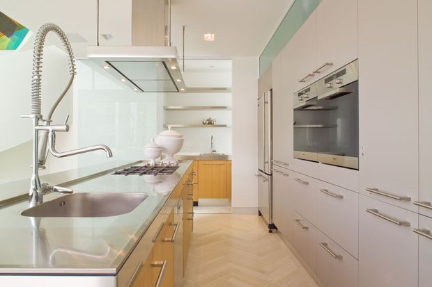 Bright, contemporary galley kitchen viewed along a long central work zone. The kitchen features pale built-in cabinetry, integrated stainless-steel appliances, a stainless-steel benchtop and sink, a timber island or lower cabinet run, open shelving, recessed task lighting, and a herringbone timber floor. A colourful abstract artwork is partially visible at the edge of the frame.