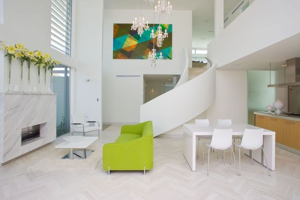 Bright, spacious open-plan contemporary interior with a double-height living and dining area. The room features a sculptural curved white staircase, white marble fireplace surround, pale herringbone flooring, full-height glazing with horizontal louvres, a vivid lime-green armchair, white dining furniture, and a compact kitchen with timber cabinetry. Multiple crystal chandeliers and colourful abstract artwork add decorative contrast.