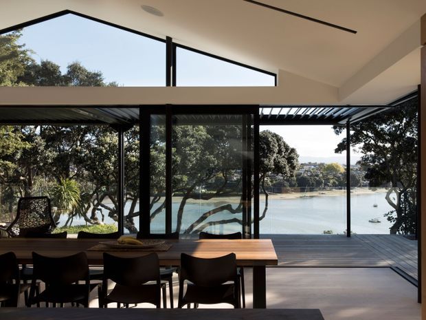 A contemporary open-plan dining and living space with a large timber dining table and dark dining chairs in the foreground. Floor-to-ceiling black-framed glazing opens directly to a timber deck and a covered outdoor area, with expansive views across a calm waterfront inlet. The room has a high sloping ceiling, clerestory glazing, minimalist recessed linear lighting, and strong indoor-outdoor connection.
