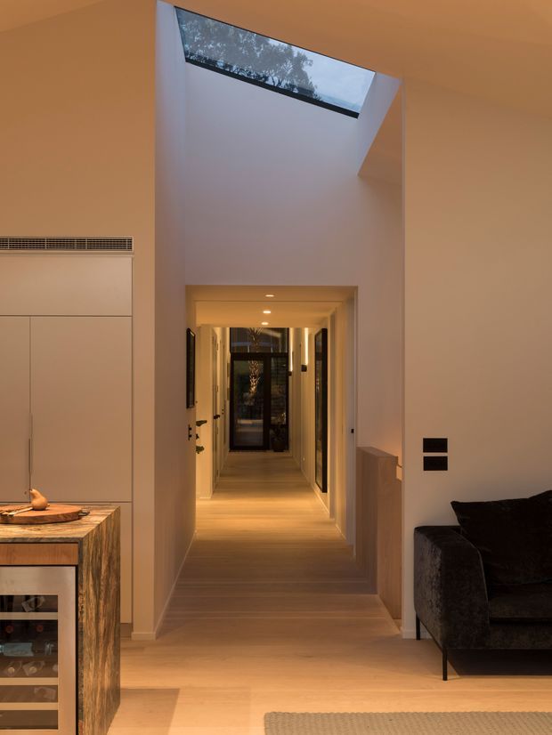 Warm-toned interior view looking from a living area down a long, minimalist hallway. Pale timber flooring, smooth light walls, recessed ceiling lights, concealed joinery, and dark-framed internal doors create a refined contemporary passage. A high angled skylight or clerestory window brings in daylight and views of trees, while partial living-room furniture and a timber-fronted cabinet are visible in the foreground.