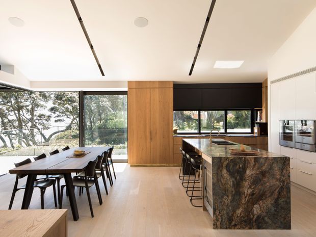 Bright, contemporary open-plan kitchen and dining area with full-height glazing, pale timber flooring, dark built-in cabinetry, a large stone island with integrated seating, and a timber-clad central storage wall. The dining table sits beside expansive windows overlooking trees and water, while linear recessed ceiling lighting and integrated appliances reinforce the clean modern design.