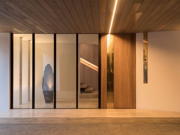 A refined contemporary interior hallway or lift lobby with a warm timber-lined ceiling and pale timber flooring. Black-framed glazed partitions reveal adjacent rooms and a stair, while a timber-clad lift door and narrow metallic control panel form a vertical feature. Linear recessed lighting provides a warm glow, and the space has a clean, minimalist architectural character.