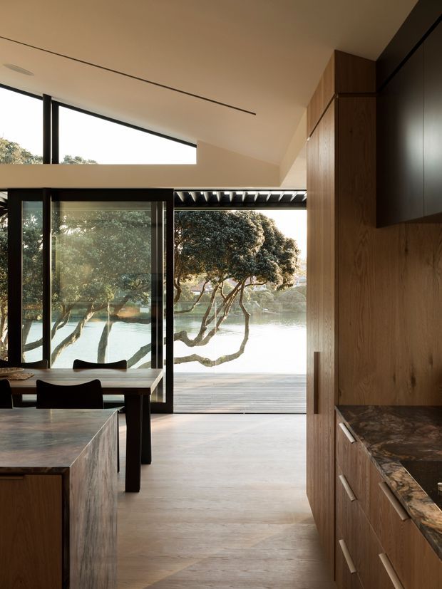 A warm, refined open-plan kitchen and dining area with extensive black-framed glazing opening toward a deck and leafy waterside outlook. The kitchen features full-height timber veneer cabinetry, a dark veined stone benchtop, and a timber-clad island. A dining table sits beside the glazing, while the minimalist ceiling includes recessed linear detailing and a projecting skylight-like opening.