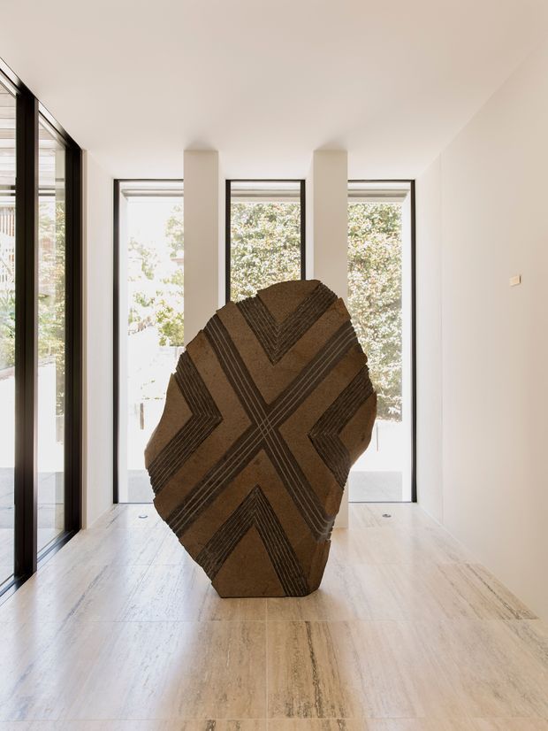 A bright, minimalist interior corridor or entry space with full-height black-framed glazing and narrow vertical windows overlooking greenery. Pale stone or travertine-style flooring and white walls create a calm gallery-like setting. A large sculptural brown geometric object with dark linear detailing stands prominently in the centre of the room.