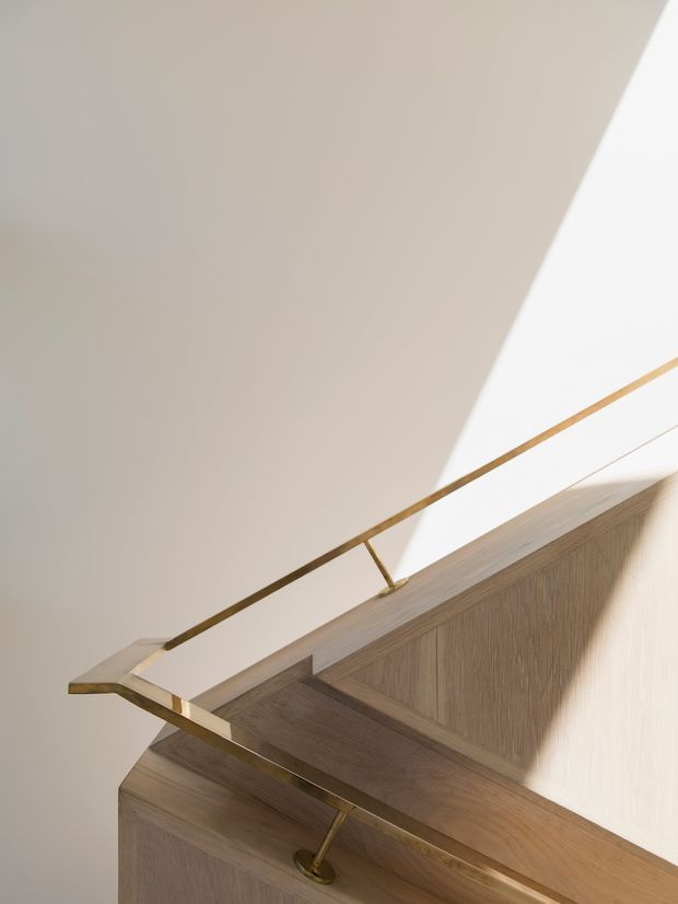Close-up architectural detail of a sculptural timber stair or landing with a slim brass/gold-toned metal balustrade and warm natural light. The image emphasises refined joinery, wood grain, geometric lines and strong angular shadows against pale walls.
