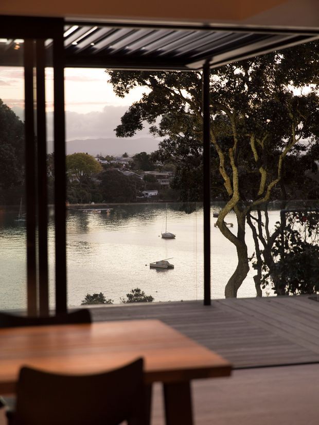 A warm, dimly lit interior dining area opens onto a timber deck with expansive floor-to-ceiling glazing overlooking a calm waterfront inlet. A timber dining table and dark chairs occupy the foreground, while a covered outdoor terrace with a slatted roof frames the view of trees, distant homes, hills, and moored boats.