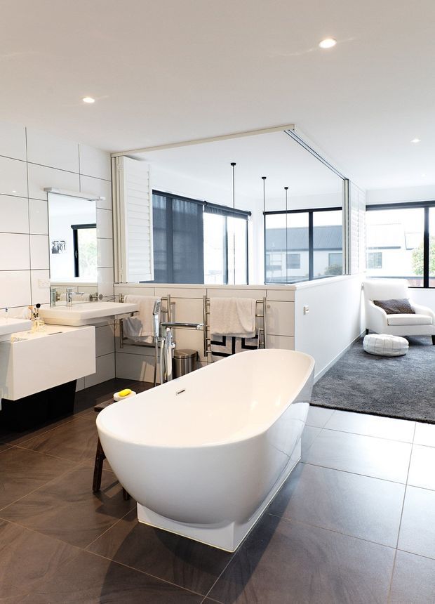Bright contemporary bathroom with a freestanding white bathtub in the foreground, twin white vanities, wall-mounted mirrors, towel rails, and large-format tiled surfaces. A glazed opening connects directly to a carpeted lounge or sitting area with an armchair, while expansive black-framed windows bring in abundant natural light.