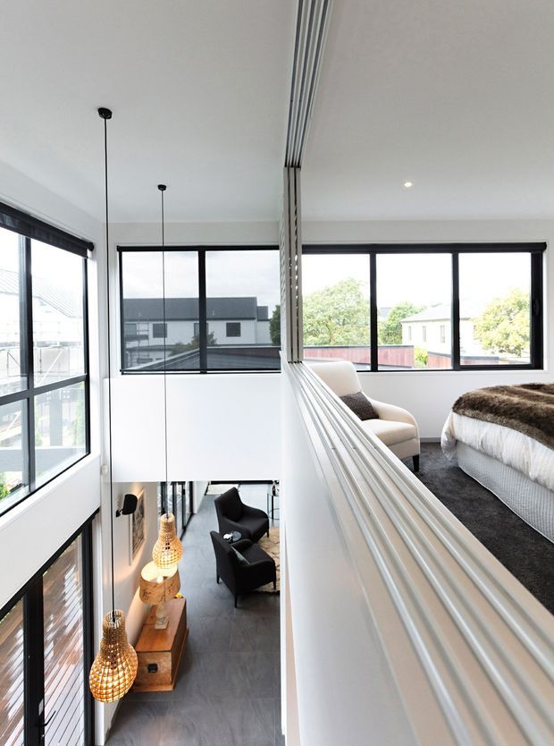 A bright contemporary upper-level bedroom overlooking a double-height living area. The room has white walls and ceiling, large black-framed windows, a grey carpet, a bed with a textured brown throw, and a light upholstered armchair. A white balustrade with vertical slatted detailing overlooks the living room below, which contains dark lounge chairs and suspended woven pendant lights. The space is airy and connected by an open void with generous glazing.