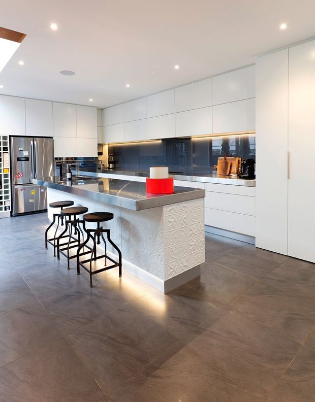 A bright, contemporary kitchen with a large central island and four black metal bar stools. The kitchen features extensive white flat-panel cabinetry, integrated appliances, a dark reflective splashback, under-cabinet lighting, a grey stone-look benchtop, and a decorative embossed tile finish on the island base. Large grey floor tiles and recessed ceiling lights complete the clean, modern space.