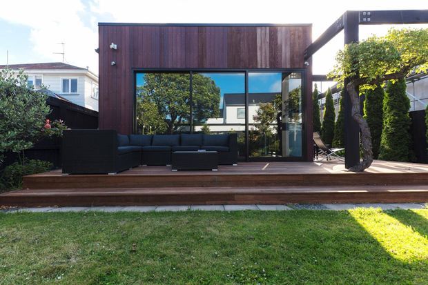 Contemporary backyard extension with dark vertical timber cladding, extensive floor-to-ceiling glazing, and a broad timber deck. The deck is furnished as an outdoor lounge with a dark sectional sofa and low coffee table, framed by a pergola and landscaped planting. Reflections reveal surrounding residential buildings and mature trees.