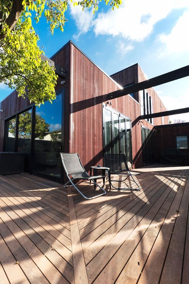 A sunlit timber deck courtyard beside a modern multi-level home clad in vertical dark-stained timber boards. Floor-to-ceiling glazing opens onto the deck, with two lounge chairs, a small side table, and a dark outdoor sofa beneath a simple overhead frame. Strong shadows, mature tree foliage, and blue sky create a warm contemporary outdoor setting.