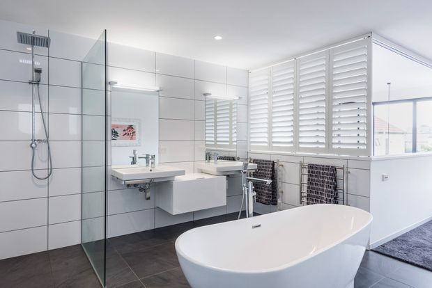 Bright contemporary bathroom with large white wall tiles, a frameless glass walk-in shower, wall-mounted vanity with integrated basin, freestanding white bathtub, plantation shutters, and dark tiled flooring. The room has a clean, high-contrast monochromatic palette and abundant natural light.