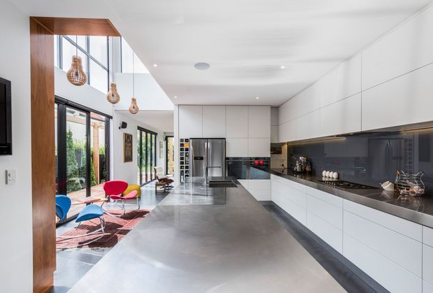 A bright, contemporary open-plan kitchen with extensive white handleless cabinetry, a long dark stone island and matching benchtop, and a dark reflective splashback. Full-height glazing and glazed doors connect the kitchen to an adjacent living area, while timber framing and woven pendant lights add warmth. Colourful lounge furniture and a patterned rug are visible beyond the kitchen.