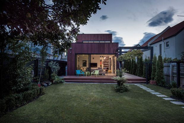 Twilight view of a compact contemporary backyard extension, with a dark timber-clad box-shaped structure opening onto a raised timber deck. The glazed opening reveals a warmly lit interior, while a manicured lawn, stepping-stone path, hedges, fencing, and neighbouring residential buildings frame the scene.