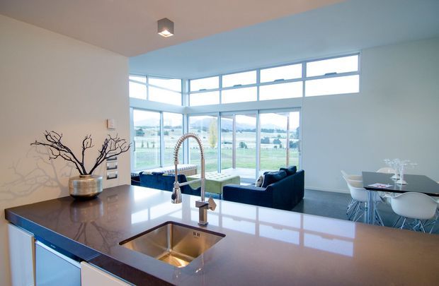 Open-plan contemporary kitchen and living area viewed from behind a large island, with a glossy dark stone benchtop, undermount sink, tall curved mixer tap, integrated cabinetry, and a dining area beyond. Floor-to-ceiling glazing and high clerestory windows provide expansive views and abundant daylight. A compact living area with a dark sofa and a modern dining setting is visible in the background.