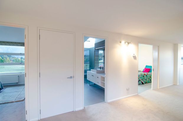 A bright, neutral hallway or landing with multiple white interior doors. A bathroom with a vanity is visible through the central doorway, while a bedroom with colourful bedding is visible through the right doorway. The left doorway opens toward a room with expansive glazing and a water view. The space has pale carpet, white painted walls, simple flush doors, and soft wall lighting.
