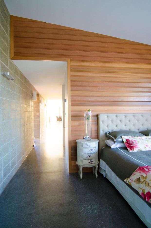 A bright bedroom with a partially visible bed, upholstered tufted headboard, floral bedding, and a small distressed bedside table. The room features a prominent horizontal timber-lined feature wall and ceiling, contrasting with a pale rendered or masonry wall and dark polished concrete-like flooring. An open doorway leads to a bright hallway.
