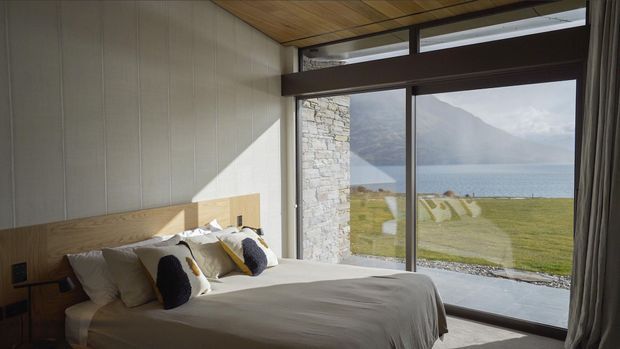 A calm, modern bedroom with a low timber headboard and neatly made bed, positioned beside full-height glazing that opens the room to a dramatic lakeside mountain view. The room features vertical light wall panelling, a timber-lined ceiling, dark window framing, a stone-clad structural wall, and soft neutral bedding with dark accent cushions.