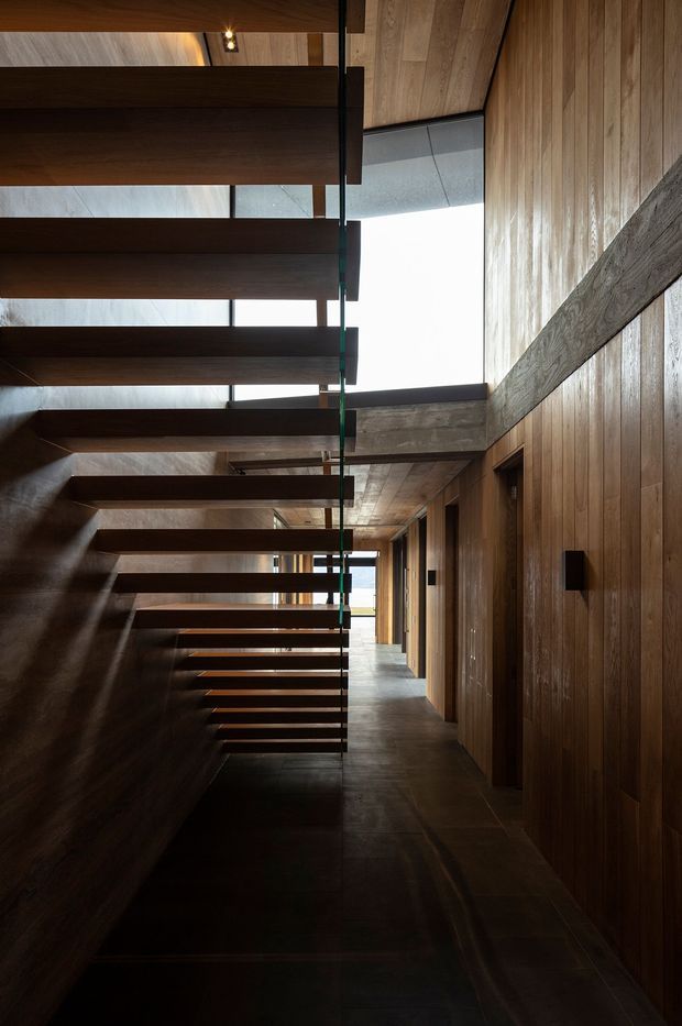 A dramatic interior hallway beside an open-riser staircase. The space features extensive dark timber wall panelling and stair treads, exposed structural beams, dark stone or concrete flooring, and large clerestory-style glazing that brings in daylight. The corridor has a warm, moody, highly crafted architectural character with strong linear geometry.