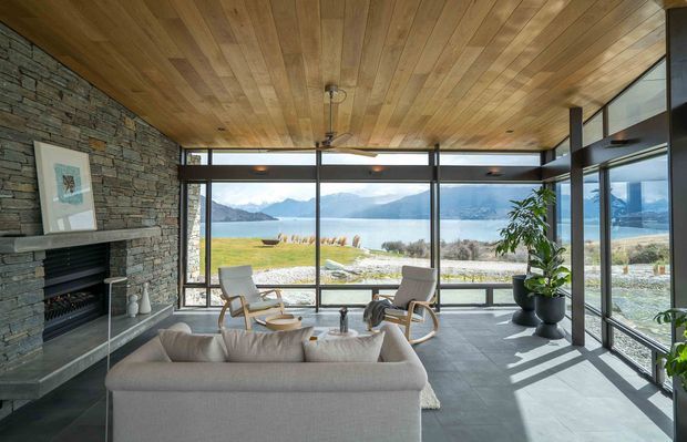A spacious contemporary living room with expansive floor-to-ceiling glazing overlooking a lake and mountains. The room features a dramatic natural stacked-stone fireplace wall, dark large-format floor tiles, extensive warm timber ceiling lining, a pale sectional sofa, lounge chairs, indoor plants, and a minimal coffee table. Black-framed glazing creates strong geometric lines and brings abundant natural light into the interior.