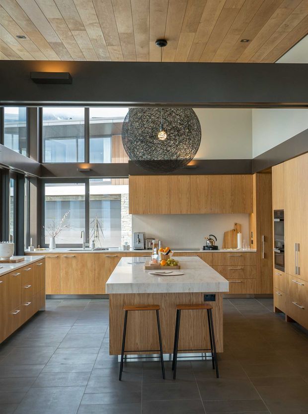 A spacious contemporary kitchen with extensive light timber cabinetry, a large pale stone island with two stools, dark grey tiled flooring, and high-level glazing that brings in abundant natural light. The ceiling is clad in warm timber boards, while dark structural beams and window frames create contrast. A large spherical woven pendant light hangs above the island, with integrated appliances and open courtyard-facing windows visible beyond.