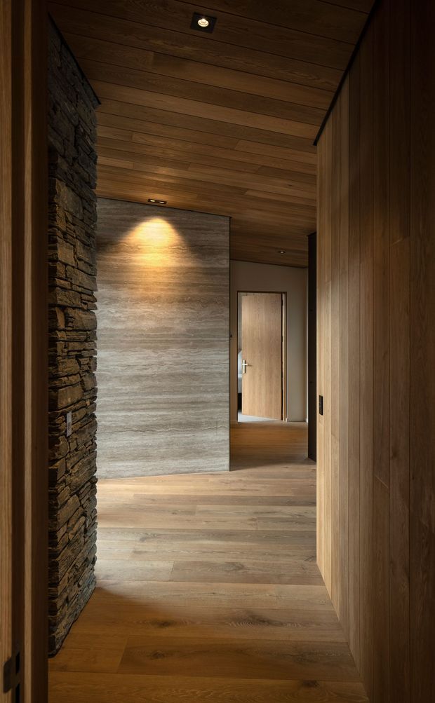 A warmly lit residential hallway or entry passage with wide-plank timber flooring and a timber-lined sloping ceiling. One wall is clad in vertical dark wood panelling, while the opposite side features a rugged stacked-stone column or wall. A softly illuminated grey stone feature wall anchors the corridor, with a timber door visible beyond. The space has recessed lighting, strong natural materials, and a refined contemporary lodge-like character.