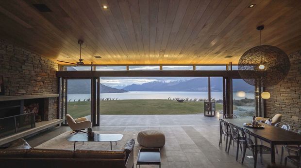 A warm, rustic-modern open-plan living and dining room with a dramatic floor-to-ceiling glazed opening overlooking a broad lake and mountainous landscape. The room features a dark stacked-stone fireplace wall, timber-lined ceiling, expansive dark-framed glazing, stone or concrete-look floor tiles, a lounge seating area, and a timber dining table beneath a large woven pendant light.