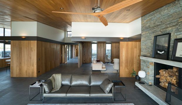 A spacious contemporary living room with a low grey leather sofa, dark tiled flooring, extensive warm timber wall and ceiling panelling, and a large stacked-stone fireplace with built-in log storage. The open-plan space extends toward a secondary seating area, with recessed lighting and a timber-bladed ceiling fan. Strong horizontal architectural lines and clerestory-like glazing create a warm, refined interior.