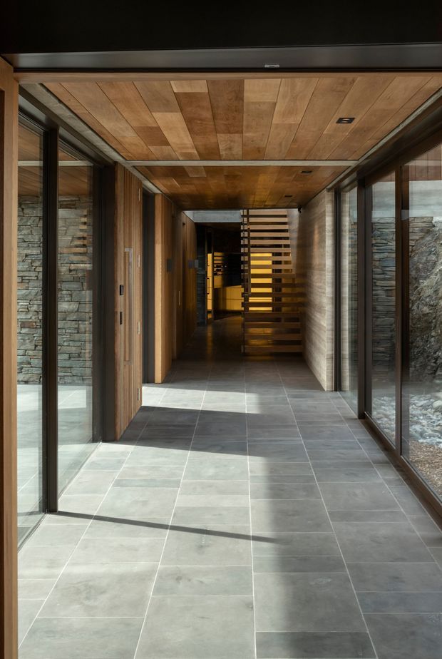 A long, contemporary interior passage or entry hall with large grey stone-look floor tiles, extensive floor-to-ceiling glazing, dark metal window and door frames, and warm timber wall and ceiling lining. Stacked stone walls are visible through the glazing, while a timber slatted screen and stair-like feature draw the eye toward the darker interior beyond. Natural light creates strong linear shadows across the floor.