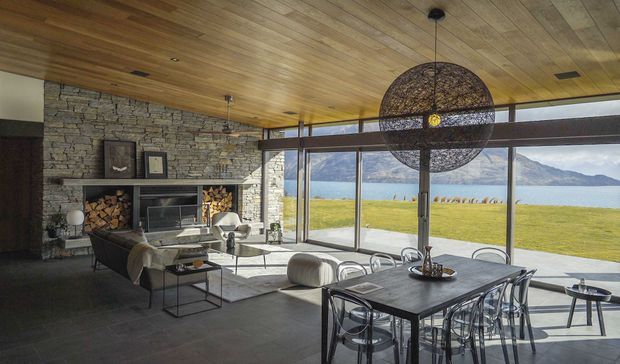 A spacious open-plan living and dining room with floor-to-ceiling glazing overlooking a lake and mountains. The room features a warm timber-lined ceiling, exposed stacked-stone fireplace wall with built-in firewood storage, dark tiled flooring, a contemporary dining table with transparent chairs, and a large spherical pendant light. The interior has a strong indoor–outdoor connection and a refined contemporary lodge-like character.