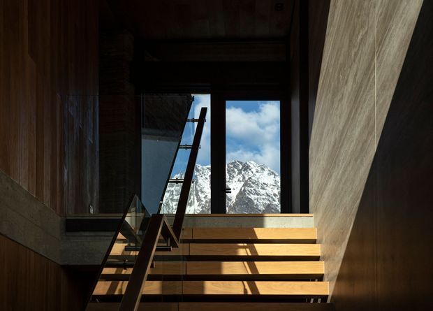 A dramatic interior view of a timber stairway ascending toward tall windows that frame snow-covered mountain peaks and blue sky. The space features dark timber wall and ceiling surfaces, warm wood stair treads, a slender dark handrail, and strong sunlight creating bold shadows. The architecture has a refined alpine, cabin-like character with a high, darkened stair hall.