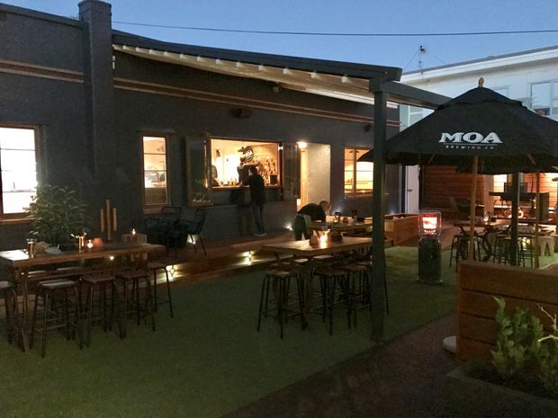 An evening view of an outdoor hospitality courtyard or beer garden, with timber communal tables, bar-height stools, umbrellas, planter boxes, and outdoor heaters. A dark rendered building with an open serving window and warm interior lighting forms the backdrop, while a lighter adjacent building is visible beyond.