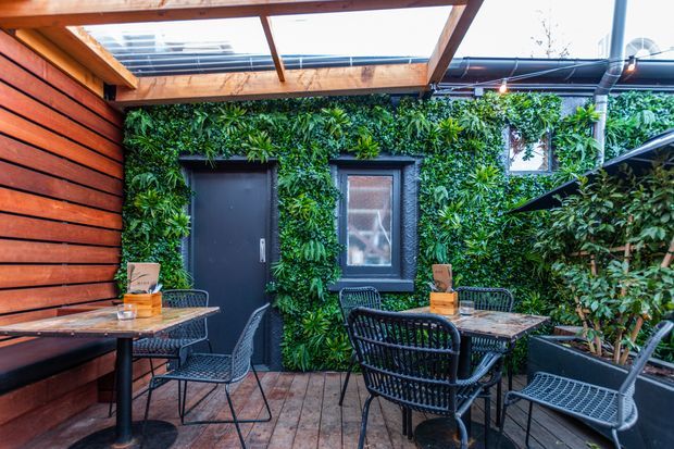 A compact outdoor dining courtyard or terrace with café-style tables and woven metal chairs. The space features a lush vertical green wall around a dark door and window, warm horizontal timber cladding, timber decking, and a timber pergola with translucent roofing overhead. The atmosphere is intimate and leafy, with a strong indoor-outdoor hospitality feel.