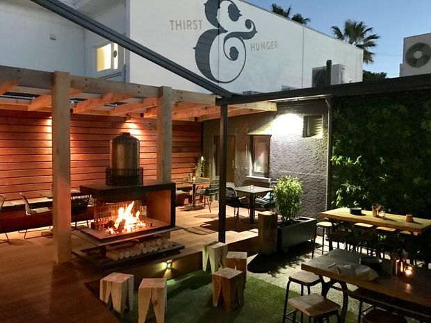 An atmospheric outdoor dining courtyard at dusk, featuring timber pergolas, warm slatted-wood walls, communal tables and stools, a prominent open fireplace, planted greenery, and a white rendered building with a large illustrated sign reading “THIRST & HUNGER.”