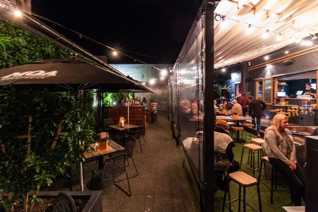 A nighttime view of a lively outdoor dining and hospitality venue. A narrow landscaped passage with café tables, chairs, umbrellas, festoon lighting and greenery runs alongside a warmly lit enclosed or semi-enclosed dining area with bar seating and patrons. The space has a casual urban atmosphere with dark finishes, timber seating and overhead lighting.