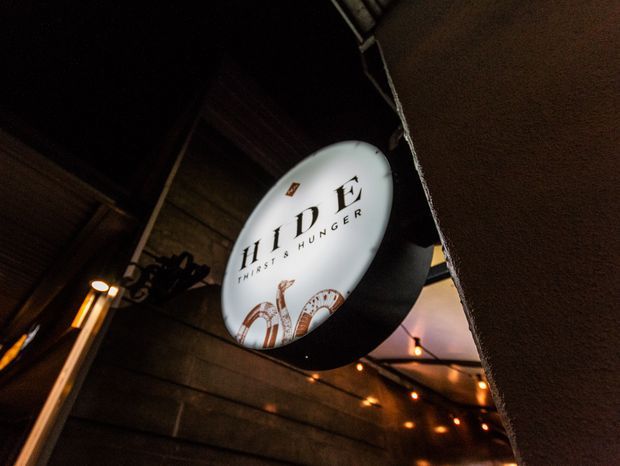 Dimly lit close-up of a round illuminated sign reading “HIDE – Fries & Hunger,” mounted against dark timber-clad walls beneath a covered structure. Warm string lights and industrial-style fixtures create an atmospheric hospitality setting.