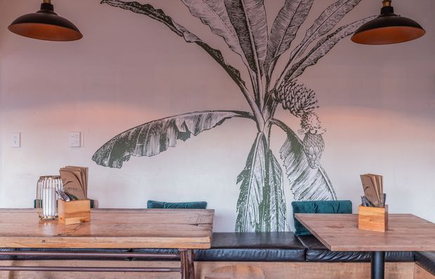 A warmly lit dining area with a long built-in black leather banquette, rustic timber tables, and teal upholstered seating. The pale wall is dominated by a large hand-drawn botanical banana plant mural, while black pendant lights hang overhead. The space has a relaxed, eclectic hospitality-style atmosphere with natural timber and dark metal details.