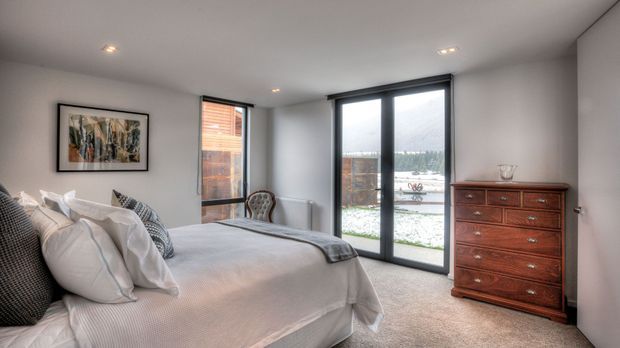A bright contemporary bedroom with a large upholstered bed, layered neutral bedding, patterned cushions, a traditional dark timber chest of drawers, framed artwork, recessed ceiling lights, and full-height glazed doors opening to an exterior area. The room has soft grey walls, pale carpet, and a calm neutral palette.