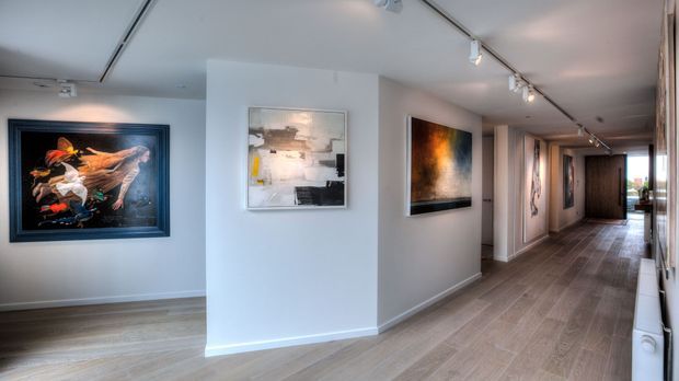 A bright, contemporary art gallery-style interior hallway with white walls, track lighting, light timber flooring, and framed artworks displayed along the corridor. The space extends toward glazed doors or windows at the far end, with a clean, minimalist exhibition-like character.