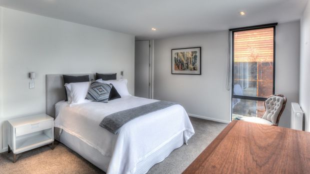 A bright, compact bedroom with a neatly made double bed, upholstered grey headboard, white bedding, patterned and dark accent cushions, and a grey throw. A small white bedside table sits beside the bed. The room has pale walls, carpeted flooring, recessed ceiling lights, framed artwork, and a tall window with timber venetian blinds. A timber desk or console with a patterned chair occupies the foreground.