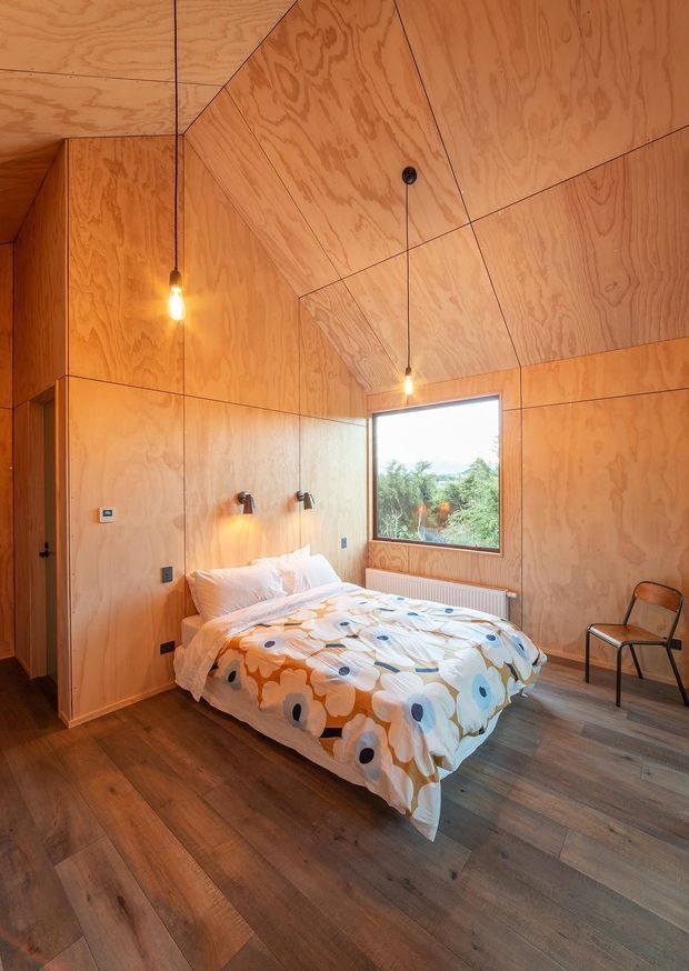 A compact bedroom clad almost entirely in warm plywood, including the walls and angled ceiling. The room features a double bed with colourful patterned bedding, wall-mounted reading lights, exposed pendant bulbs, timber flooring, a large window, built-in storage, and a simple chair. The faceted plywood lining creates a warm, cabin-like atmosphere.