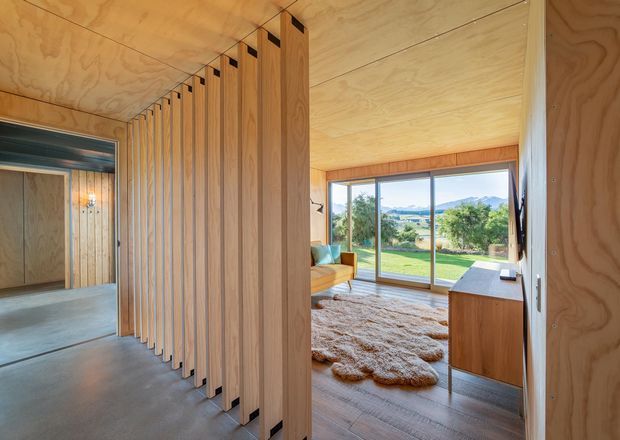 Interior view of a warm, timber-lined modern home with a full-height vertical timber slat partition separating a concrete-floored circulation area from a cozy living room. The living space has timber flooring, a low timber cabinet, textured rug, sofa, and large sliding glass doors opening to a green landscape and distant hills.