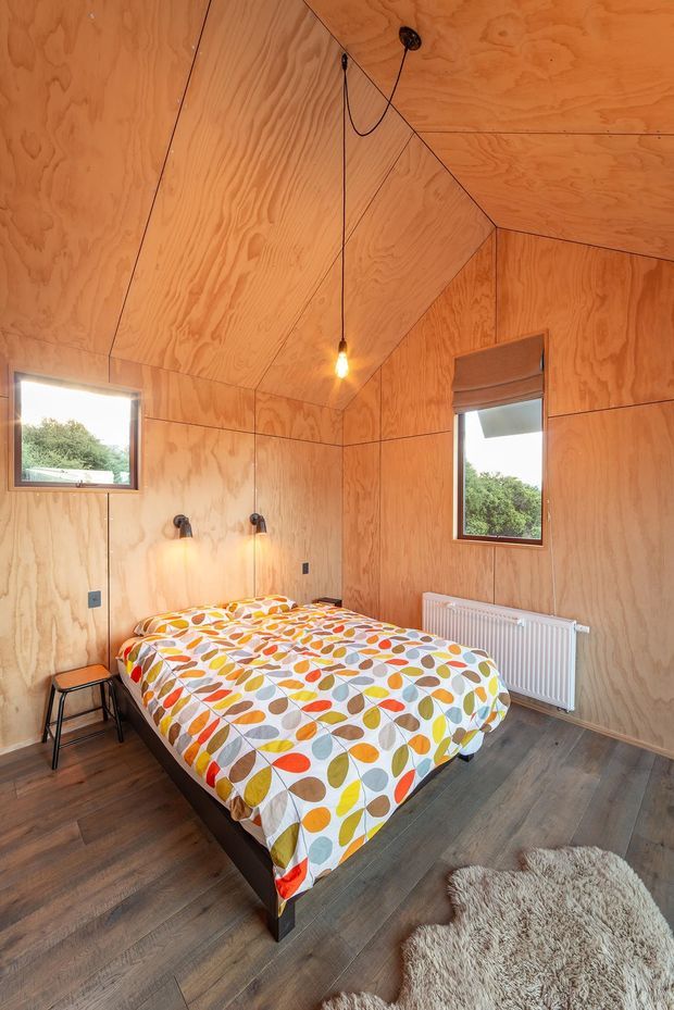 A compact bedroom clad in warm, exposed plywood panels across the walls and angular ceiling. The room contains a double bed with colourful retro leaf-patterned bedding, dark timber-look flooring, small wall-mounted reading lights, two windows with greenery visible outside, a white radiator, and a shaggy cream rug. The faceted plywood-lined ceiling creates a distinctive cabin-like interior.