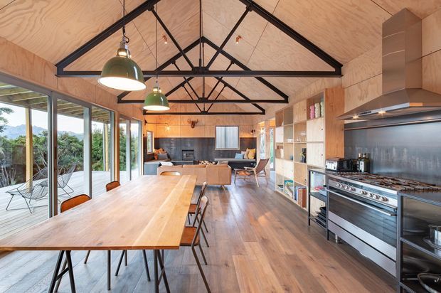 Open-plan kitchen, dining, and living space with extensive timber plywood wall and ceiling lining, exposed black steel roof trusses, timber flooring, and floor-to-ceiling glazing opening to a planted outdoor area. A large timber dining table occupies the foreground, while the kitchen features full-height timber cabinetry, a dark backsplash, stainless-steel appliances, and a substantial freestanding range cooker. The rear includes a compact living area with a sofa and armchairs.