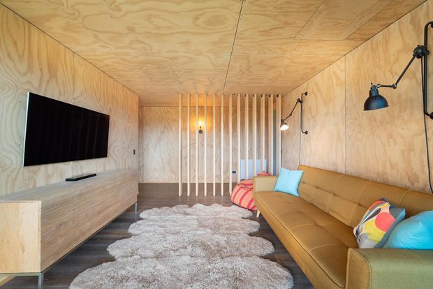 A compact, warmly lit media lounge lined entirely in pale plywood, with a wall-mounted television, low timber entertainment cabinet, long tan sectional sofa, colourful cushions, shag rug, and exposed black wall-mounted lighting. Vertical timber slats partially screen the rear of the room, creating a cosy contemporary retreat.