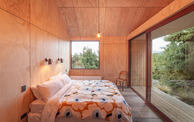 Compact bedroom lined throughout with warm plywood panels on the walls and ceiling. A double bed with patterned bedding sits against the left wall beneath two wall-mounted reading lights, while a large glazed sliding door on the right opens visually to a timber deck and landscaped greenery. A timber chair and narrow vertical timber detailing reinforce the cabin-like, natural-material character.