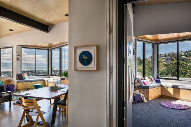 A bright contemporary open-plan interior photographed through a central partition, showing a dining area on the left and a cozy window-seat room on the right. Both spaces feature extensive black-framed corner glazing with views toward a hilly coastal setting. The dining area has a light timber table, mixed seating, pale cabinetry, and a warm timber-lined ceiling with exposed dark structural beams. The adjacent room includes built-in timber window seating, deep blue carpet, colourful cushions, and a circular patterned floor cushion. Neutral plaster walls, black metal framing, and abundant daylight create a relaxed, contemporary atmosphere.
